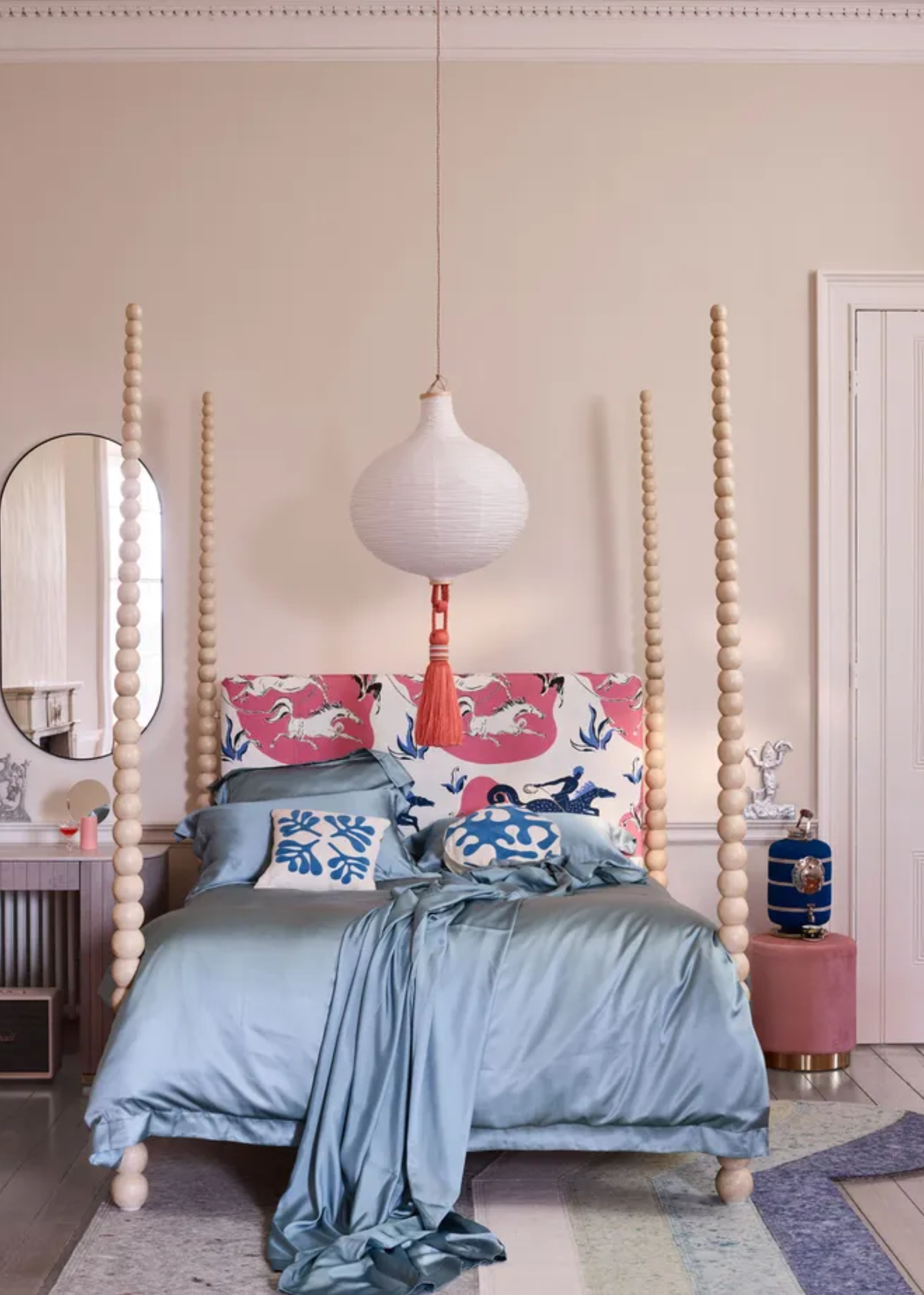 A light bedroom with bobbin-style bed posts, a patterned headboard, a low pendant rice paper lamp, and a geometric striped rug