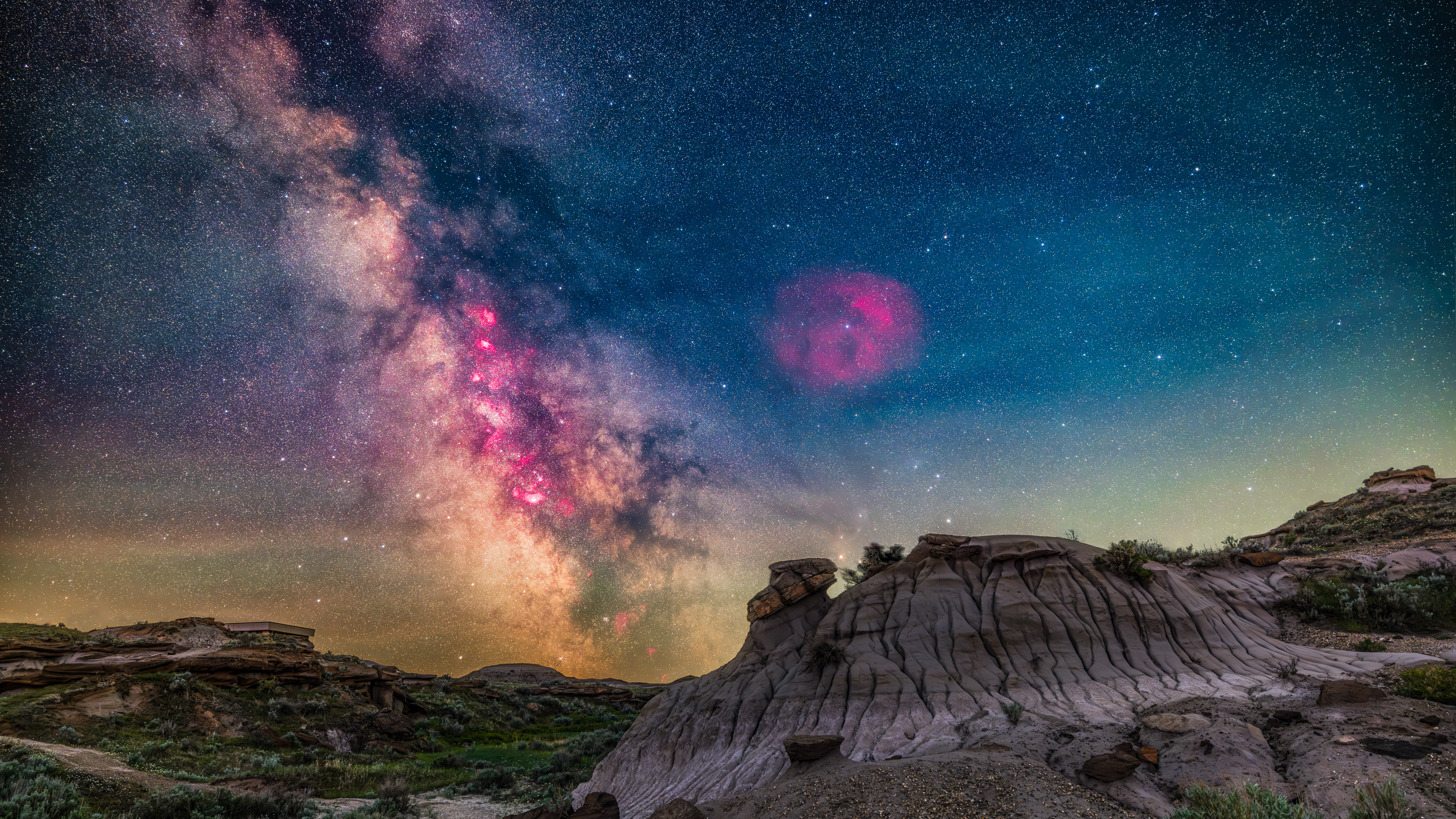 The Milky Way and its core region in Sagittarius and Scorpius is here low over the Badlands landscape of Dinosaur Provincial Park, Alberta. This was the night of May 31/June 1, 2024, when from this latitude of 50&amp;deg; 45' N the sky is not fully dark even in the middle of the night, here about 2:30 a.m. MDT. So the sky retains a blue tint. Adding to the sky colours are bands of green oxygen airglow and perhaps yellow sodium airglow. Plus some light pollution from nearby Brooks, Alberta. The mass of stars toward the galactic centre also glow with a combined yellow light, in part due to absorption of shorter wavelengths of starlight by interstellar dust in the spiral arms of the Galaxy. But the most striking colors are the red and magenta from glowing hydrogen gas in star-forming nebulae toward the galactic core. I emphasized those through the use of a filter that isolates that red wavelength of 'hydrogen-alpha.' (Photo by: Alan Dyer/VWPics/Universal Images Group via Getty Images)