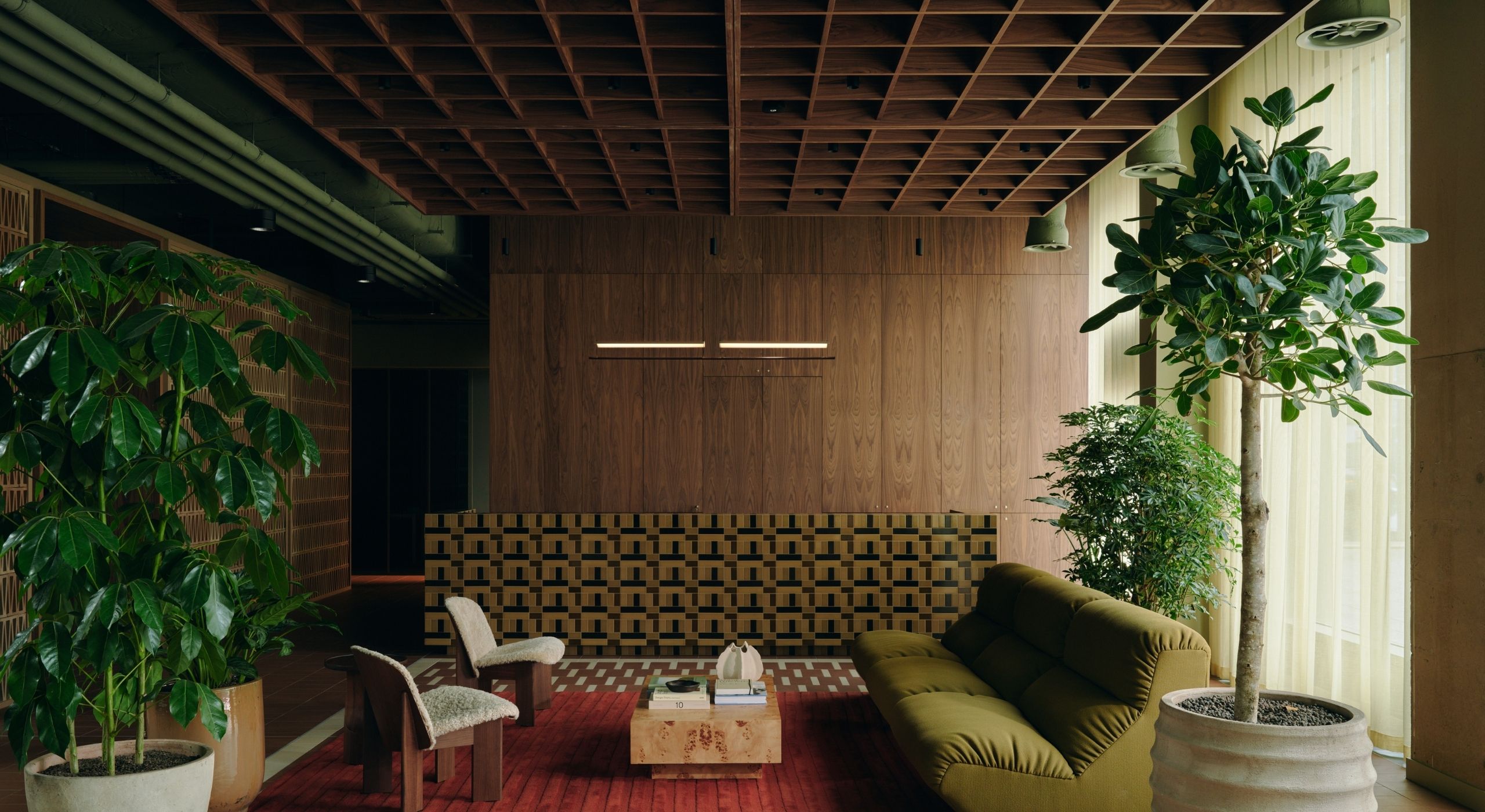 A frontal photograph of a lounge decorated with wood and upholstered seats, two olive green, oversized sofas, one checkered one color-blocked one, standing plants sitting in ceramic vases, and see-through curtains.