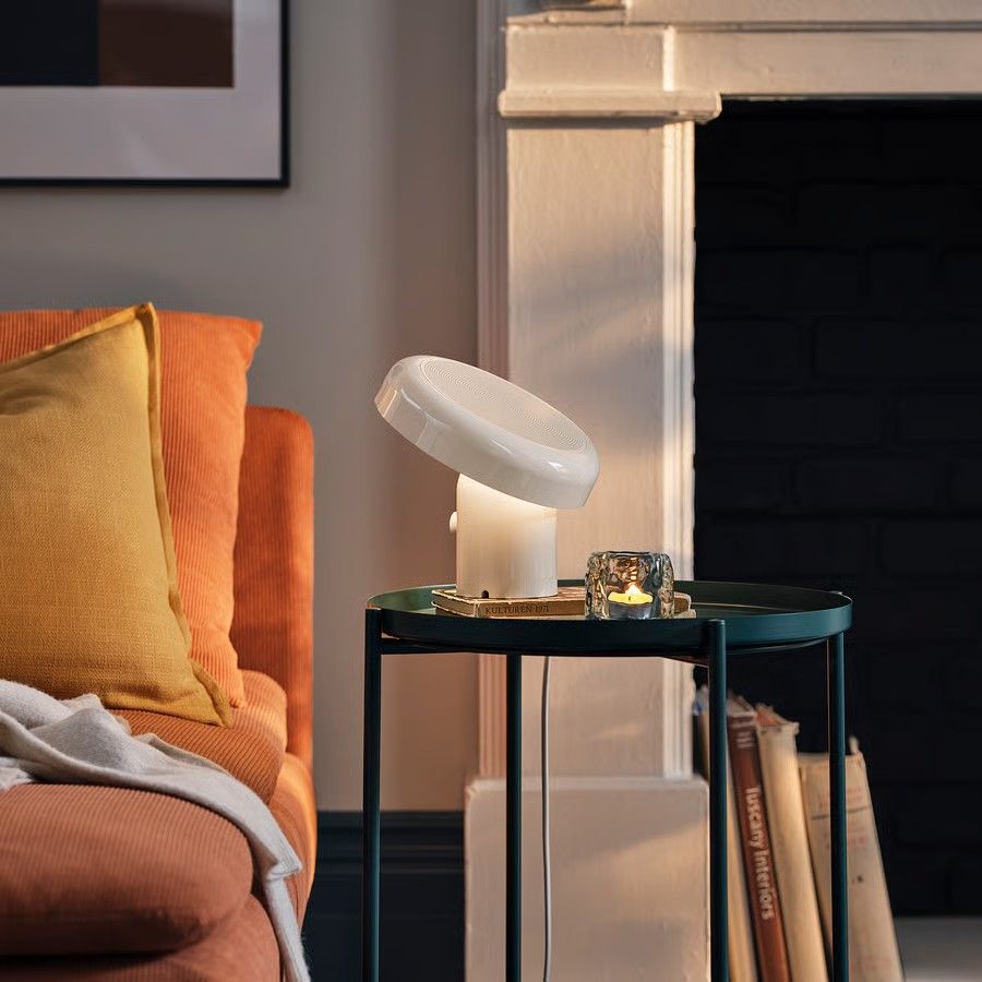 A white Bluetooth speaker lamp on a thin, green, metal side table next to an orange sofa. 