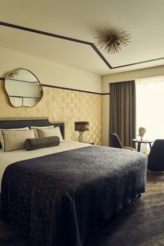 art deco-style bedroom with navy and white bed linen, dark carpets, wall panelling, spiked ceiling pendant light, bolster pillow on bed and organic-shaped mirror over bed