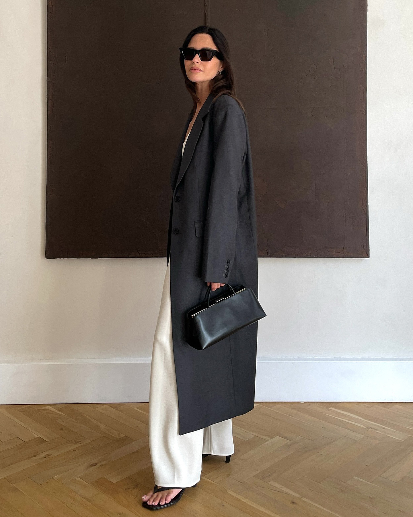 Finnish style influencer Johanna Piispa poses in black sunglasses, a long gray coat, white satin pants, heeled flip-flop sandals, a structure black bag.