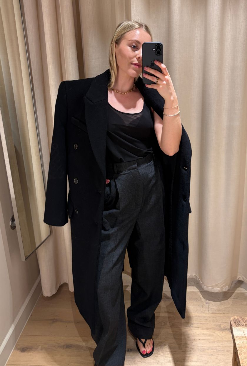 Woman wears black coat, sheer top and grey tailored trousers