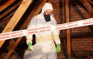 man in protective white suit and gloves wearing mask stood in loft behind warning asbestos removal tape
