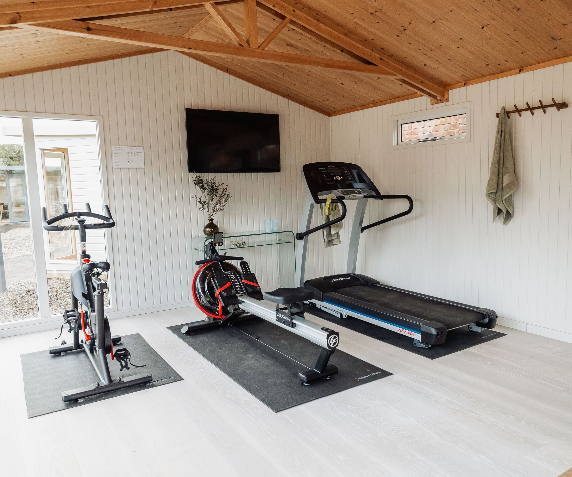 Home gym with treadmill, rowing machine and exercise bike under wooden vaulted ceiling, wall-mounted TV.