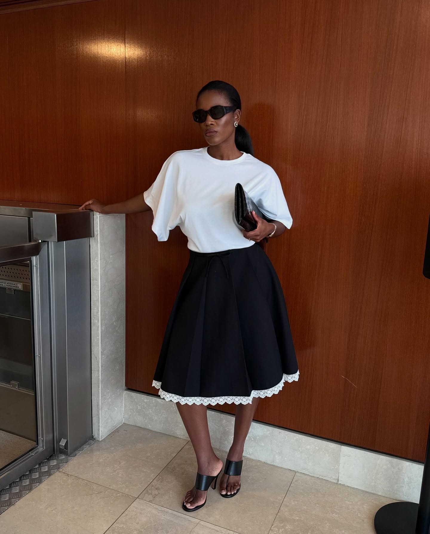 @nlmarilyn wearing a white T-shirt, black skirt, and stretch thong sandals.