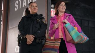 From left to right: Carra Patterson holding her hands in front of her and smiling as Kaya while standing next to Carrie Preston's Elsbeth who is holding three bags and looking to her right while smiling. 