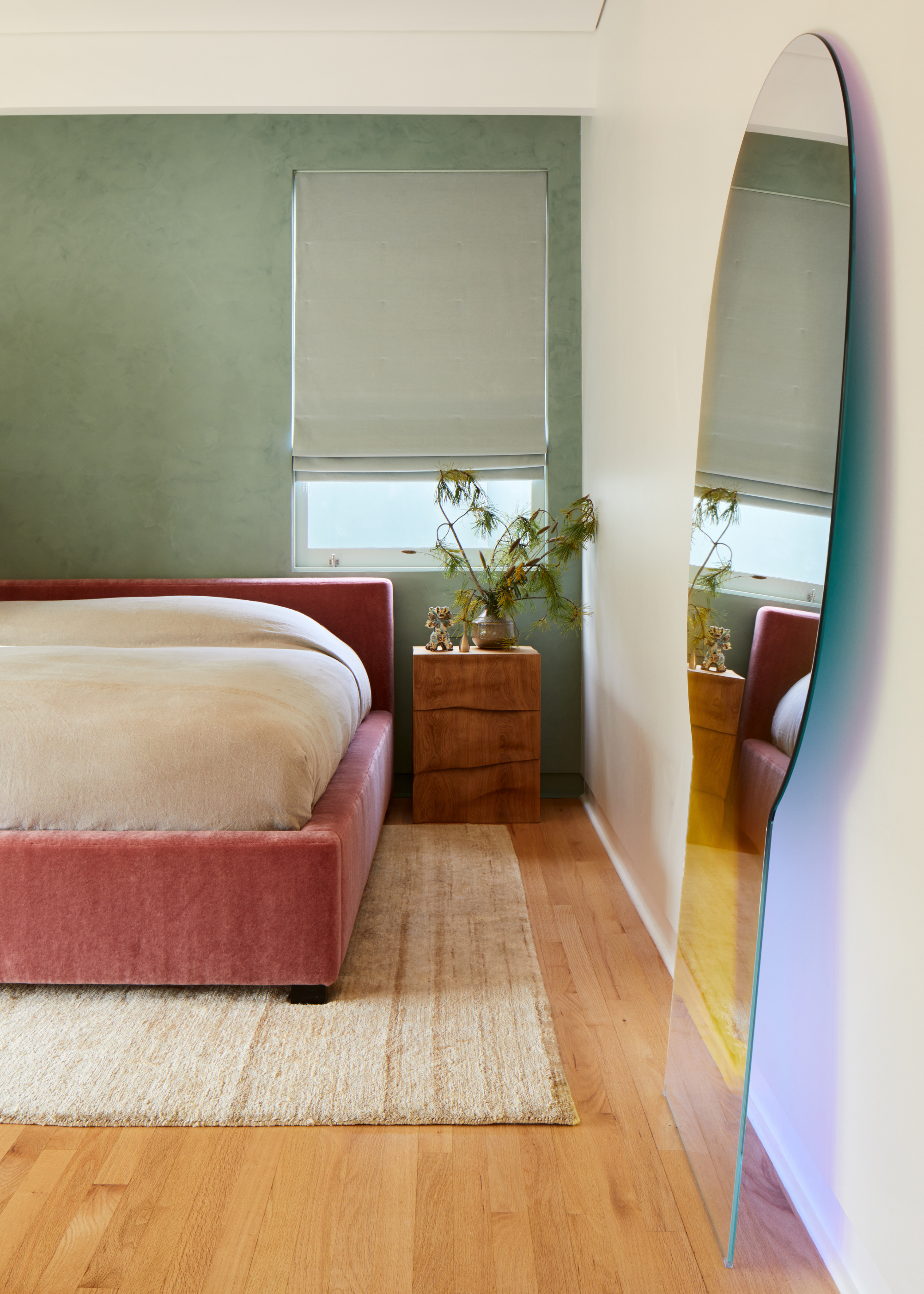 A white bedroom with a green accent wall, wood flooring, a pink bed frame, a textured rug, a wood side table with a vase of stems, and a holographic mirror on the floor