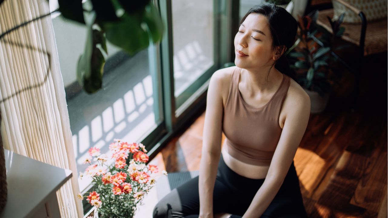 A peaceful-looking young woman sits next to sliding glass doors and a pot of flowers.