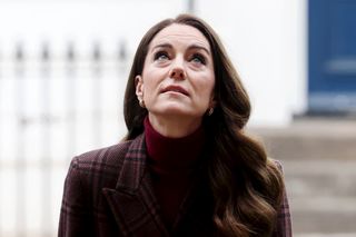 Kate Middleton looking up and wearing a plaid coat with a blue door behind her and fence