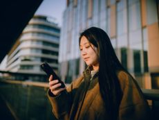 Young woman using smartphone on bridge near modern glass office buildings at sunset 