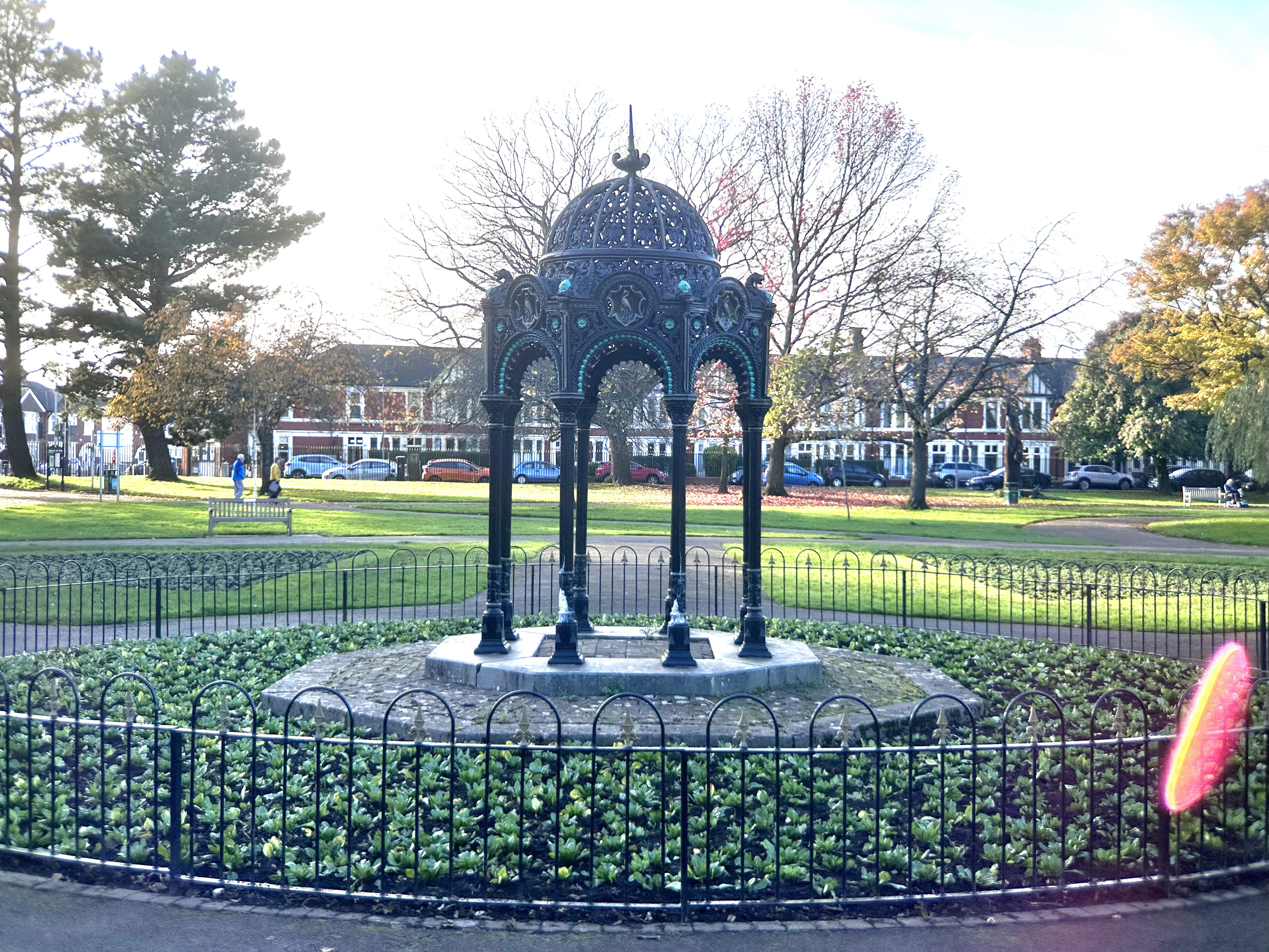 A photo of a decorative bandstand in a park on a sunner day, with a red lens flare in the lower right corner of the photo.