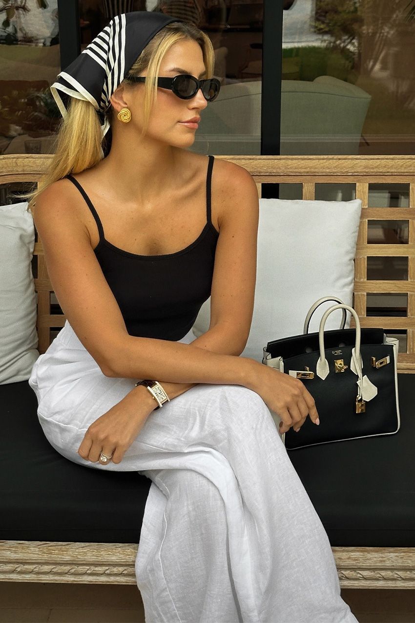 European vacation outfit idea is shown in a photo of a woman sitting outside on a bench wearing a black and white headscarf, black oval sunglasses, gold earrings, a black spaghetti strap tank top, white linen trousers, a black watch, a black and white handbag, and black flip flops