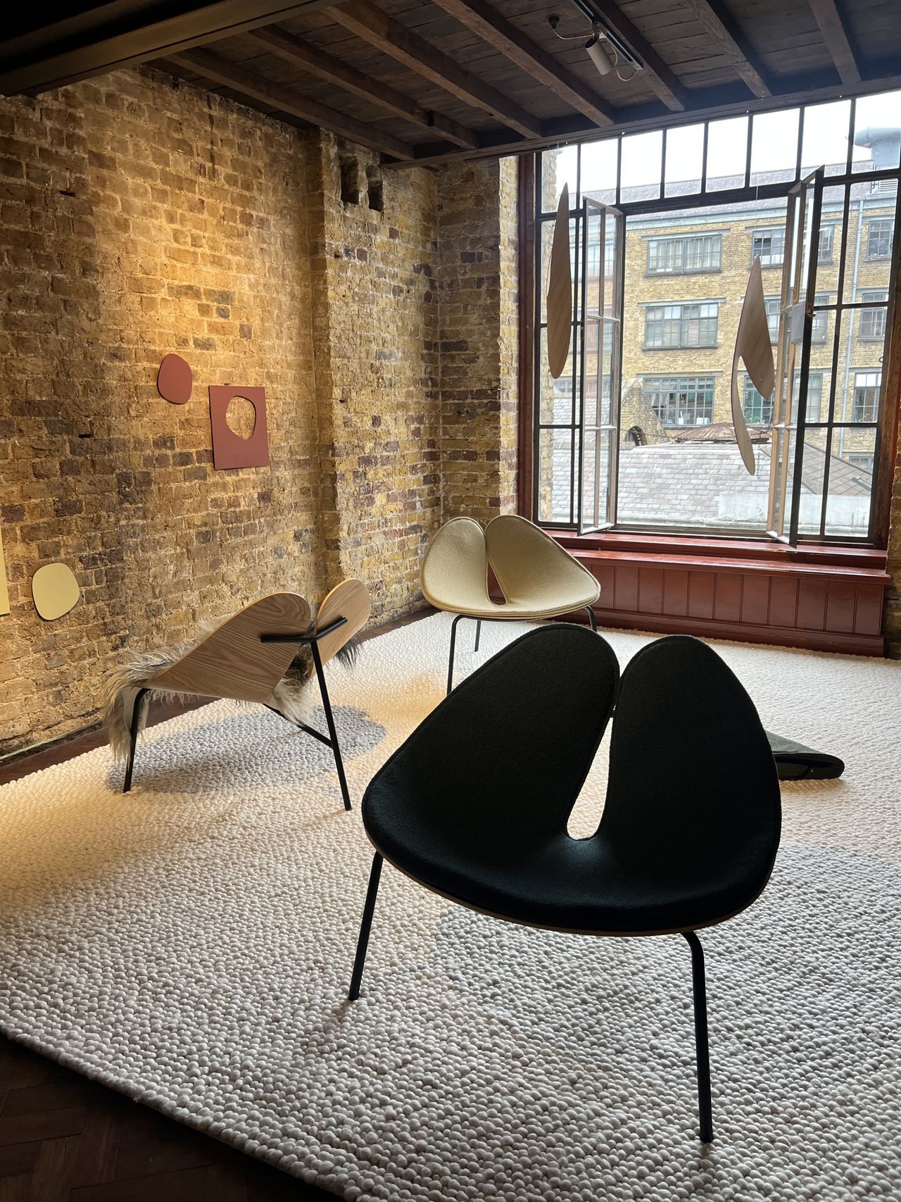 Image of three abstract side chairs arranged in a brick room with a large window on the back wall and a white, textured rug.