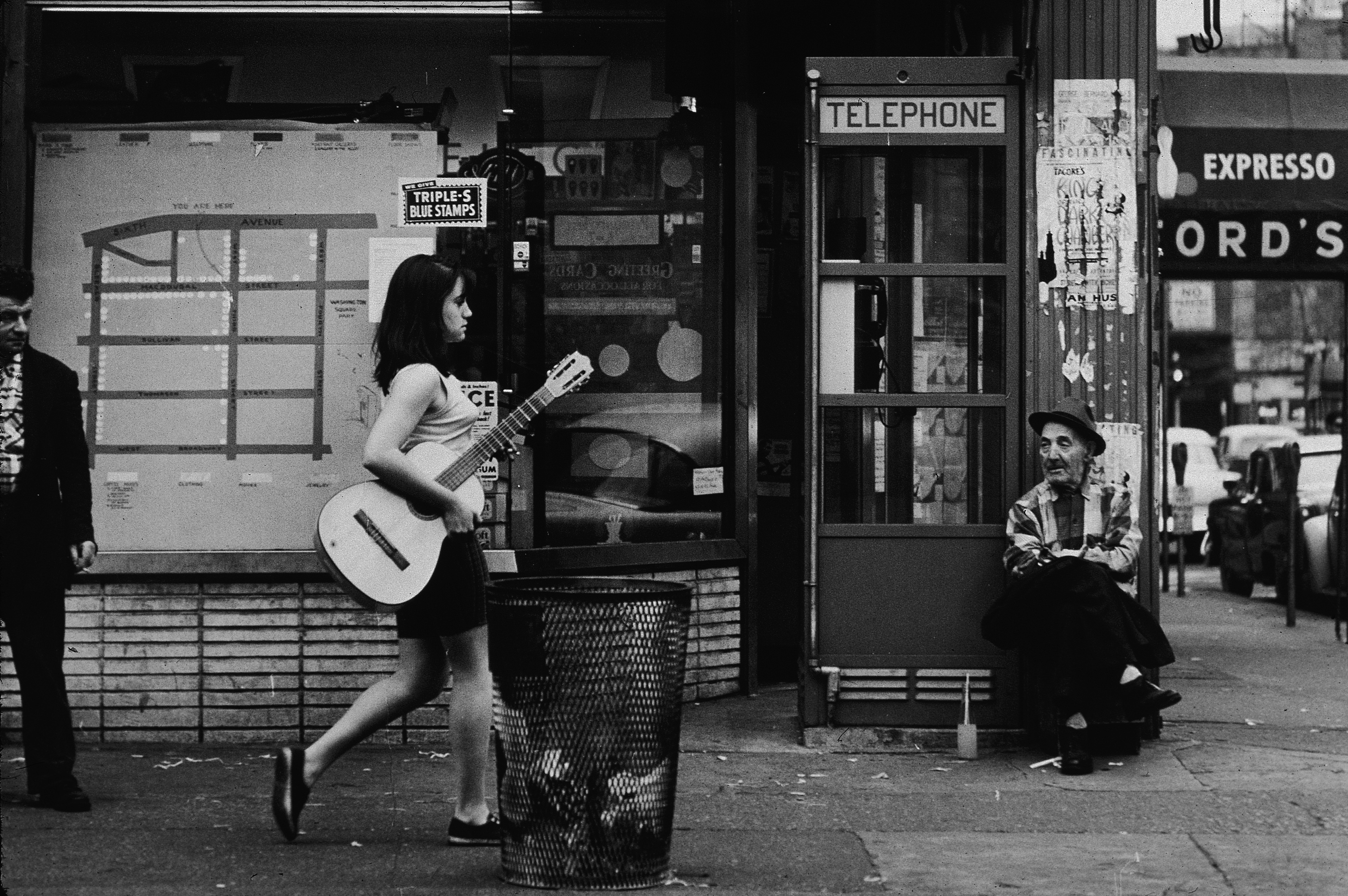 A young woman walks down a sidewalk in Greenwich Village, carrying an acoustic guitar