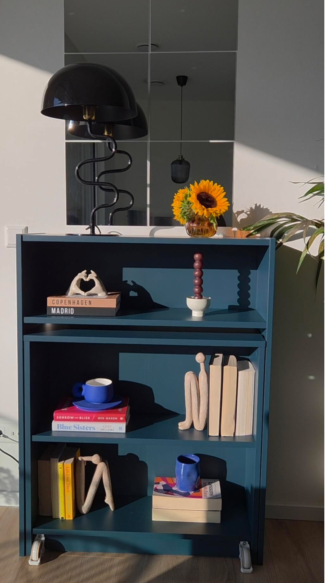 A blue-grey bookcase sits in a sun-kissed room. Various pieces of decor cover the shelves.
