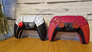 Two DualSense controllers with thumbsticks and grips attached on a wooden surface with a white brick background