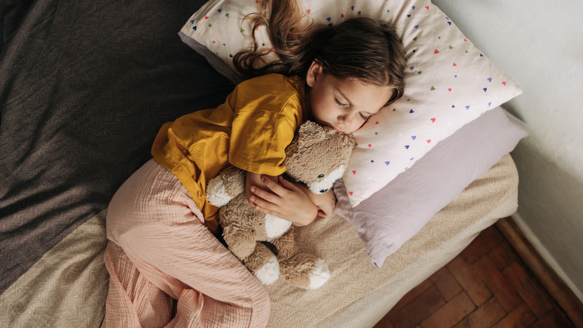 A young girl lies in bed asleep, she is lying on her side cuddling a brown and white teddy bear.