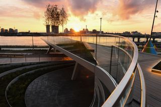 View from the walkway of The Tide riverside park and the river as the sun sets. The park features trees, greenery and colourful waterdrop-shaped structures. There are multiple buildings across the river