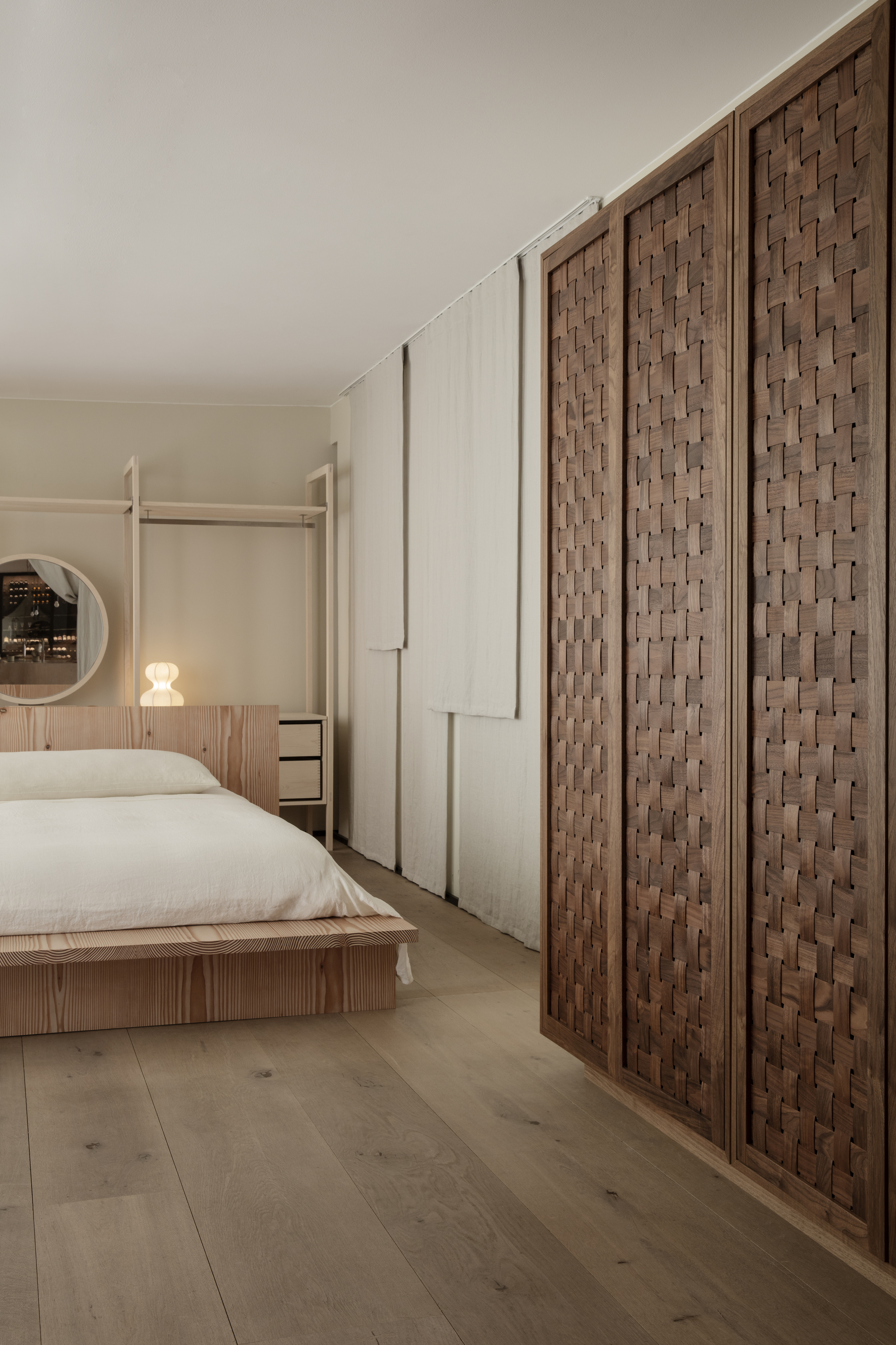 Image of a minimalist bedroom with a low, wooden bed that has white bedding. There is a large wooden built-in closet with basket weave patterned doors.