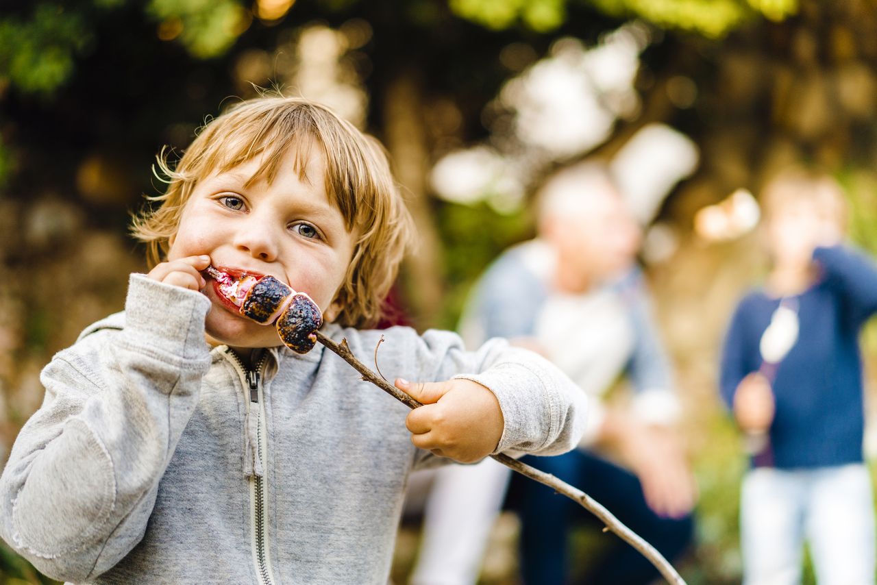 Best kids&#039; hoodies - photo of cute boy eating roasted marshmallows. Portrait of male child eating sweet food at camping. Family is in background at forest.