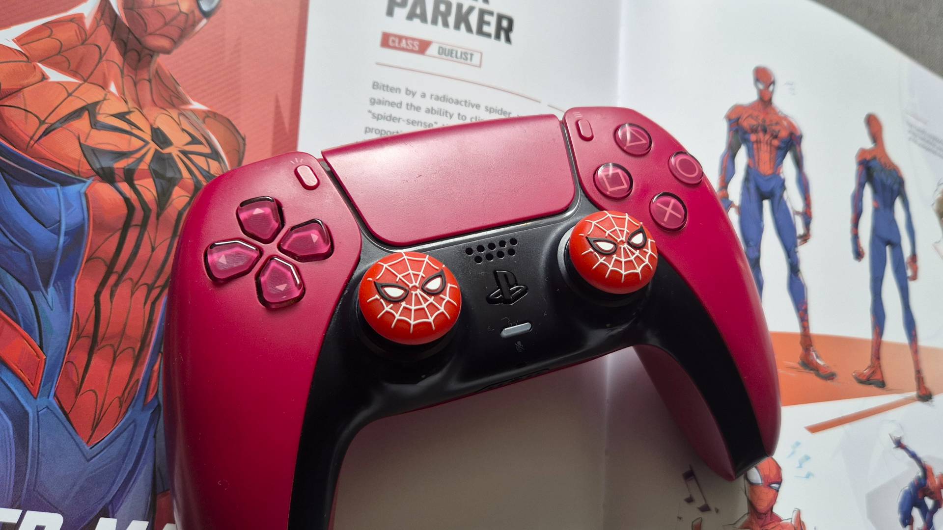 Photo by Rosalie Newcombe of a red PS5 DualSense controller with Spider-Man stick grips attached sitting on the open page showing of the art of Spide-Man in the Art of Marvel Rivals artbook.