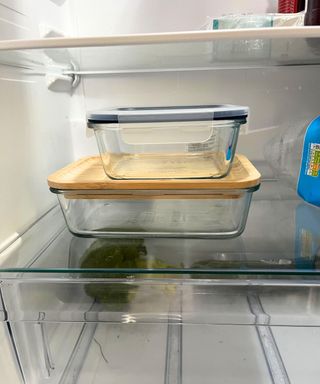 Two glass food containers, one with a bamboo lid, one with a plastic lid, stacked on top of one another in a fridge.