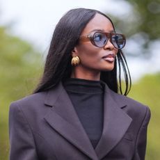 woman with super glowy skin wearing black blazer, gold earrings and tinted sunglasses
