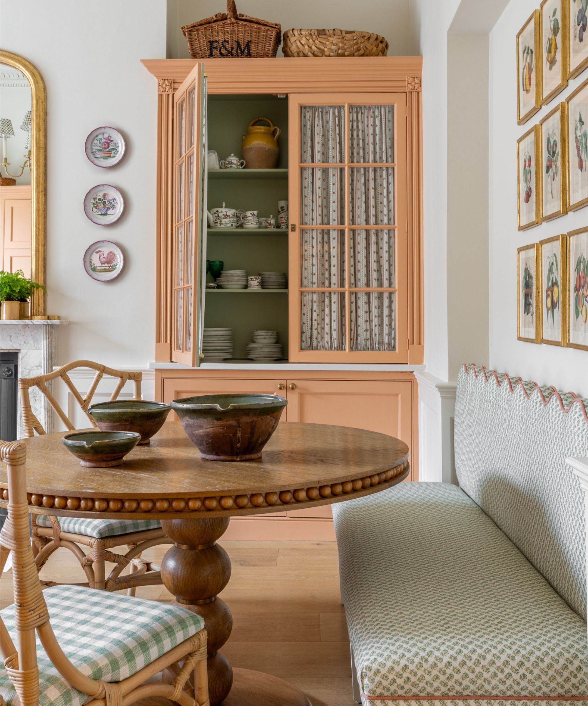 A dining area within a kitchen, with an upholstered bench seat with a scalloped edge behind a round dining table. A large pantry behind it in a light coral colour is half open showing various items of crockery inside.