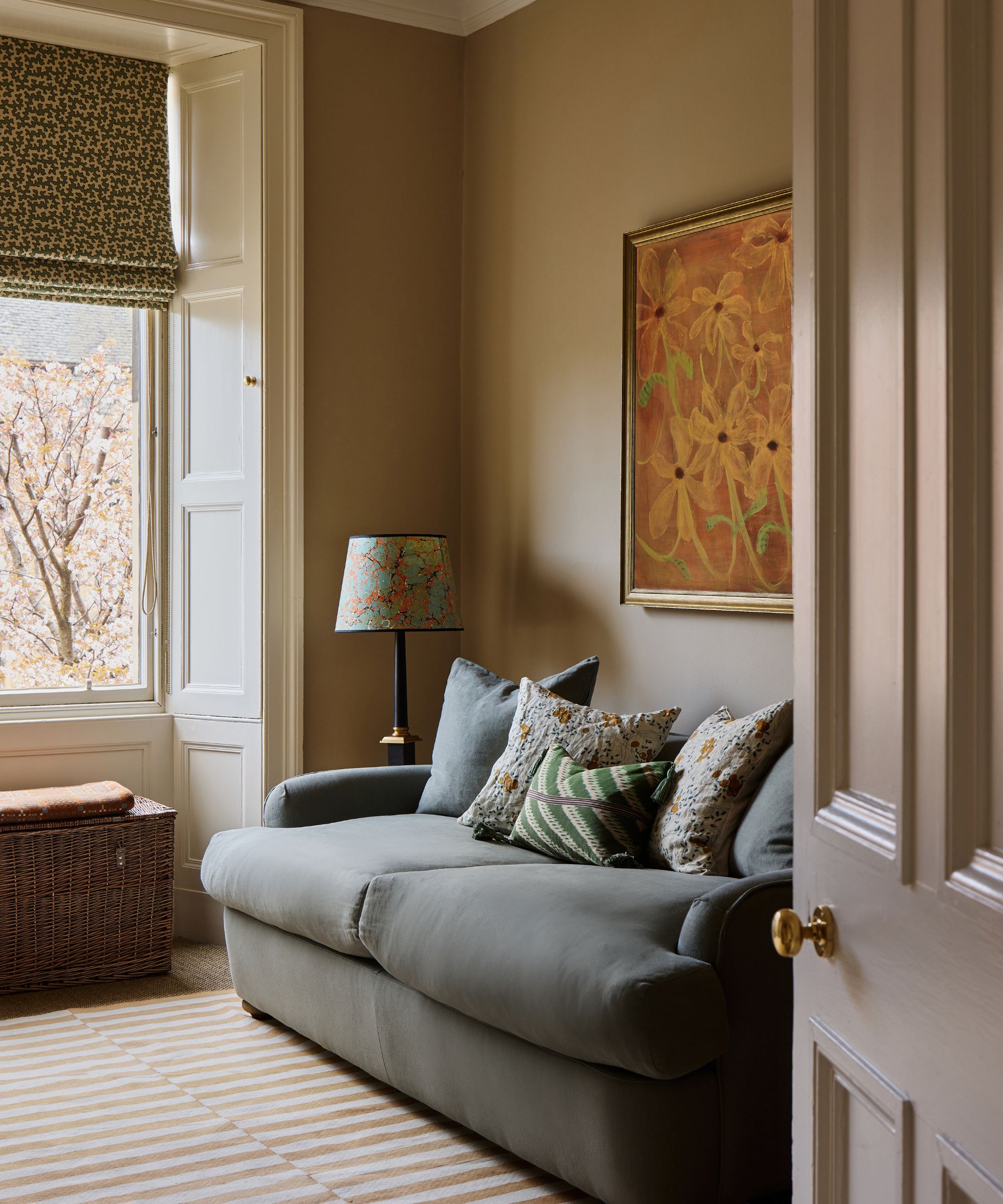 A cozy sitting room featuring a soft blue-grey sofa with floral cushions, warm ochre walls, and a large framed yellow floral painting
