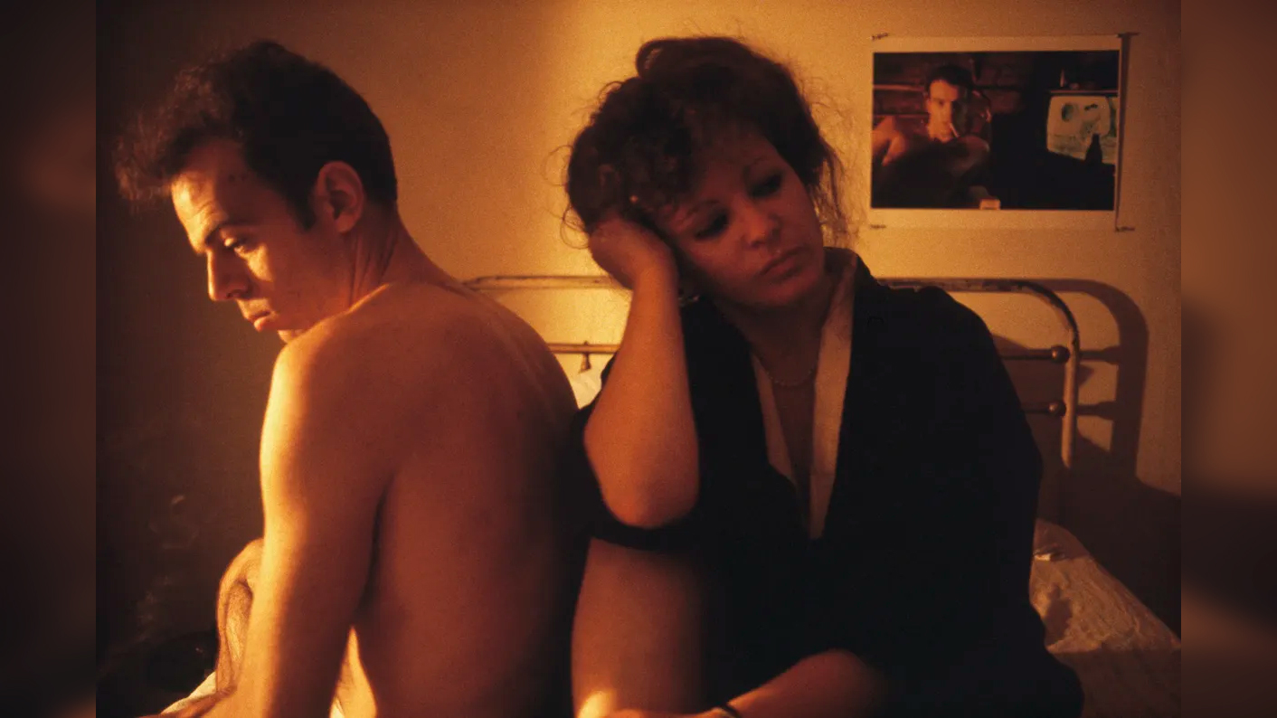 A shirtless man and a woman in a dark robe sit back to back on a bed in warm, low light, both looking downward and lost in thought.