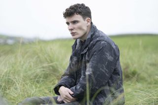 Connor Cairns (Nicholas Nunn) sits in a field of tall grass looking pensive, with his hands clasped in his lap. He is dressed all in black and grey.