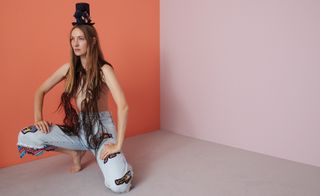 A woman on one knee wearing a pair of frayed jeans, no shirt and a black hat in front of an orange wall.