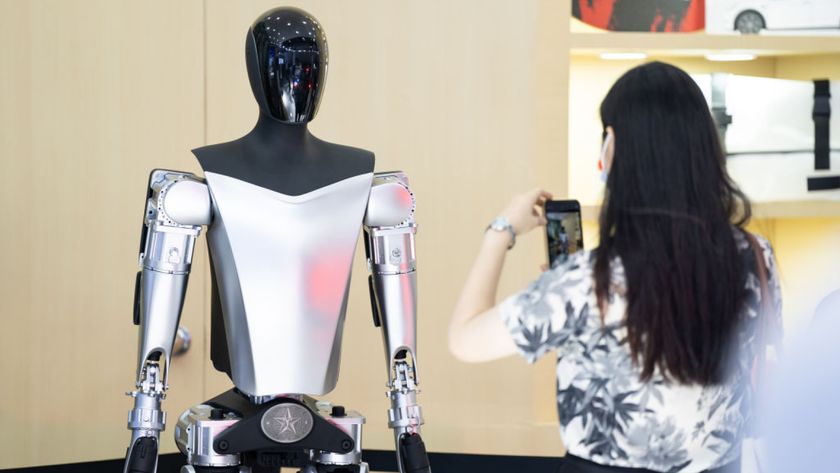 In the image a silver, faceless, humanoid, robot stands still as a lady take a photo of the robot on her phone