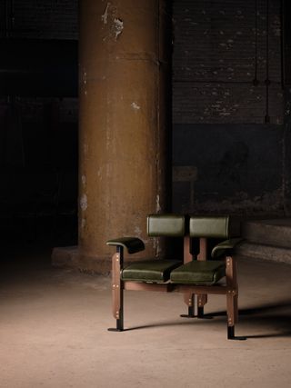 A walnut wood, steel, and brass chair, with olive green leather seating and back. It's set in an industrial factory setting.