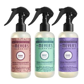 Three matching plastic bottles of room sprays with black lids, one with a pink label, one with a green label, and one with a purple label