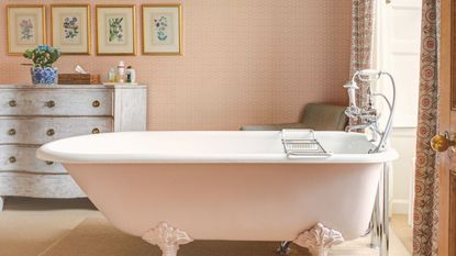 A pink wallpapered bathroom with a rolltop free standing tub. A distressed dresser in the background with eight gold framed pictures on the wall above it.