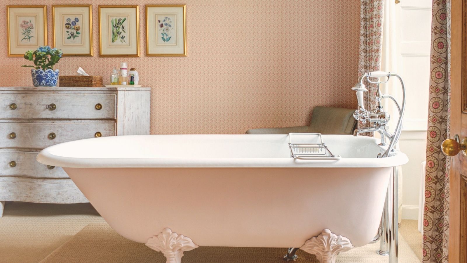 A pink wallpapered bathroom with a rolltop free standing tub. A distressed dresser in the background with eight gold framed pictures on the wall above it. 