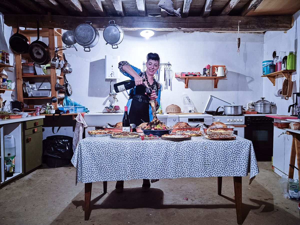 A home kitchen featuring a table laden with various baked goods, while a person pours wine, surrounded by cooking utensils and colorful kitchen items