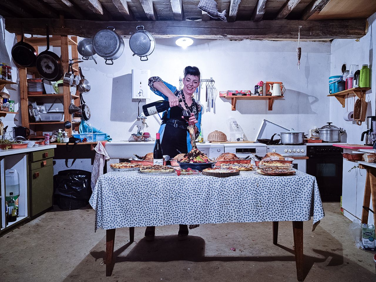 A home kitchen featuring a table laden with various baked goods, while a person pours wine, surrounded by cooking utensils and colorful kitchen items