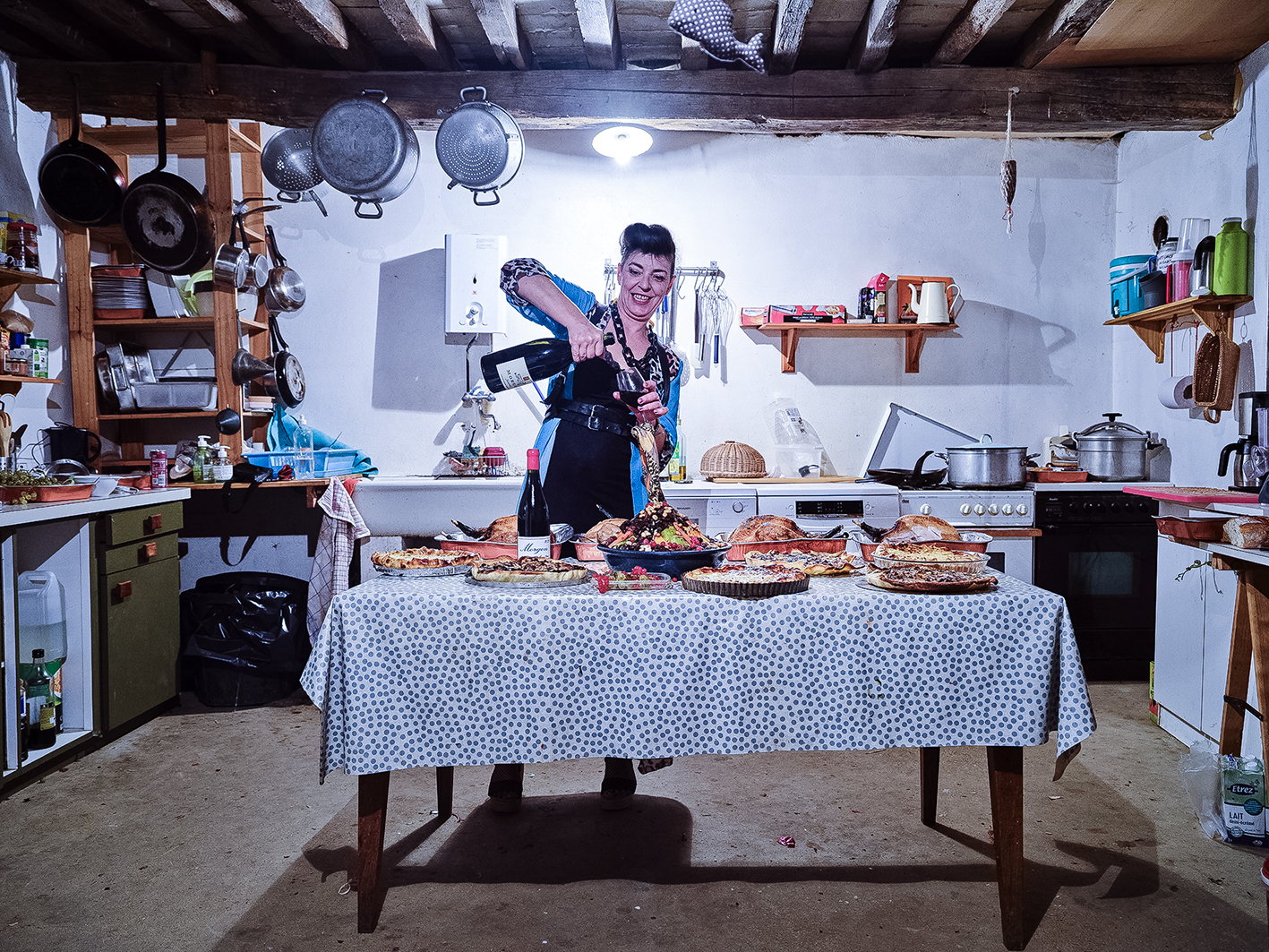 A home kitchen featuring a table laden with various baked goods, while a person pours wine, surrounded by cooking utensils and colorful kitchen items