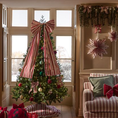 A living room with a striped sofa decorated for Christmas with a Christmas tree by the window decorated with a big striped bow tree topper and a matching tree skirt