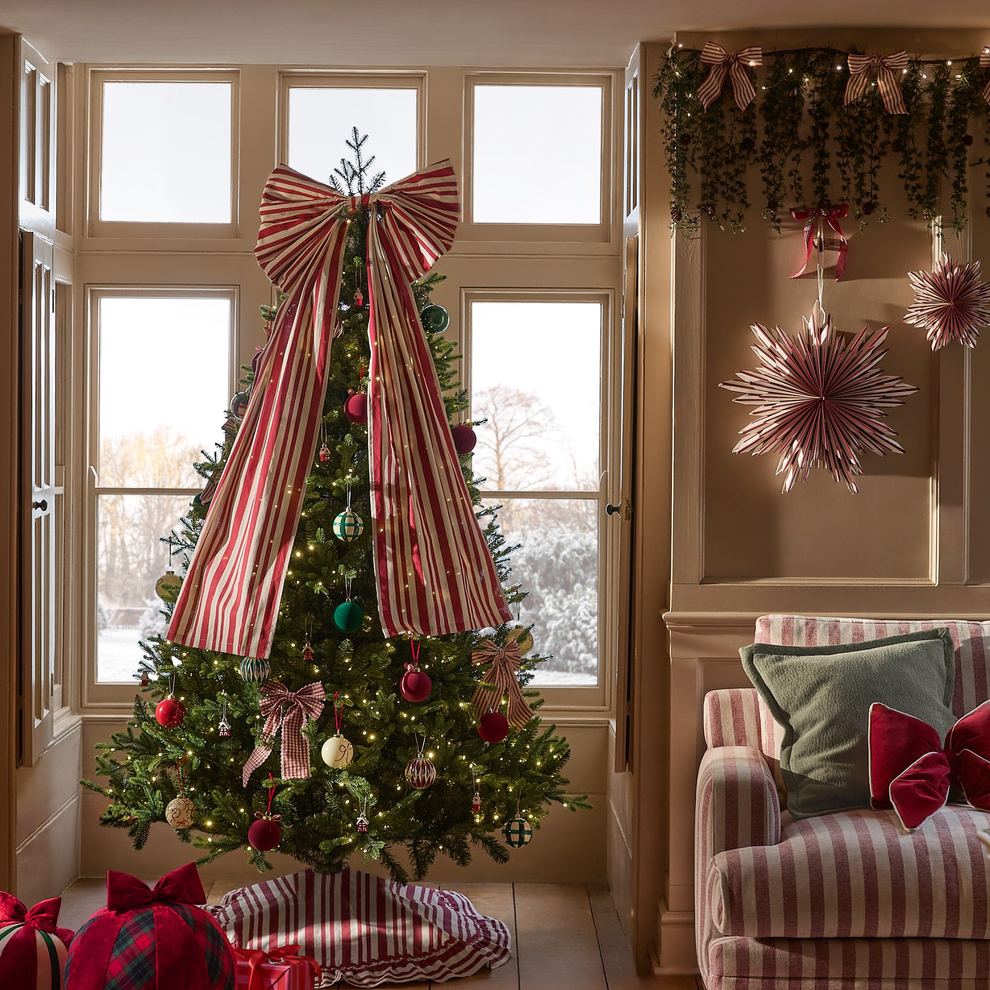 A living room with a striped sofa decorated for Christmas with a Christmas tree by the window decorated with a big striped bow tree topper and a matching tree skirt