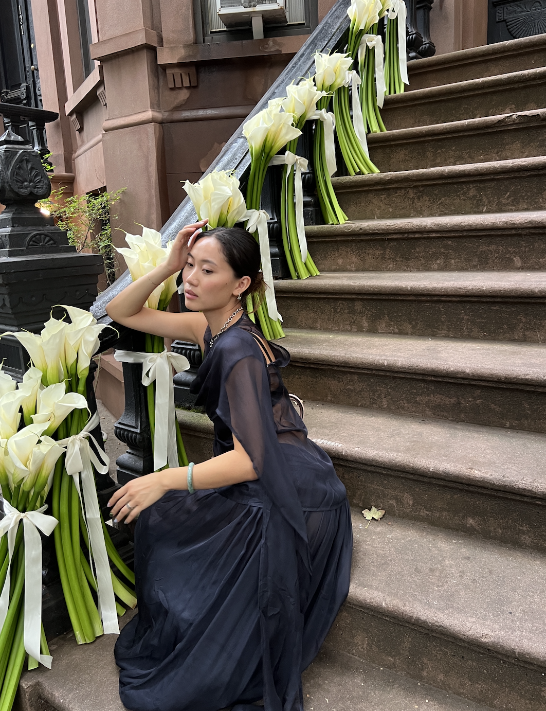 Vivian Li wearing a blue sheer maxi dress with a sash sitting on steps outside in New York City