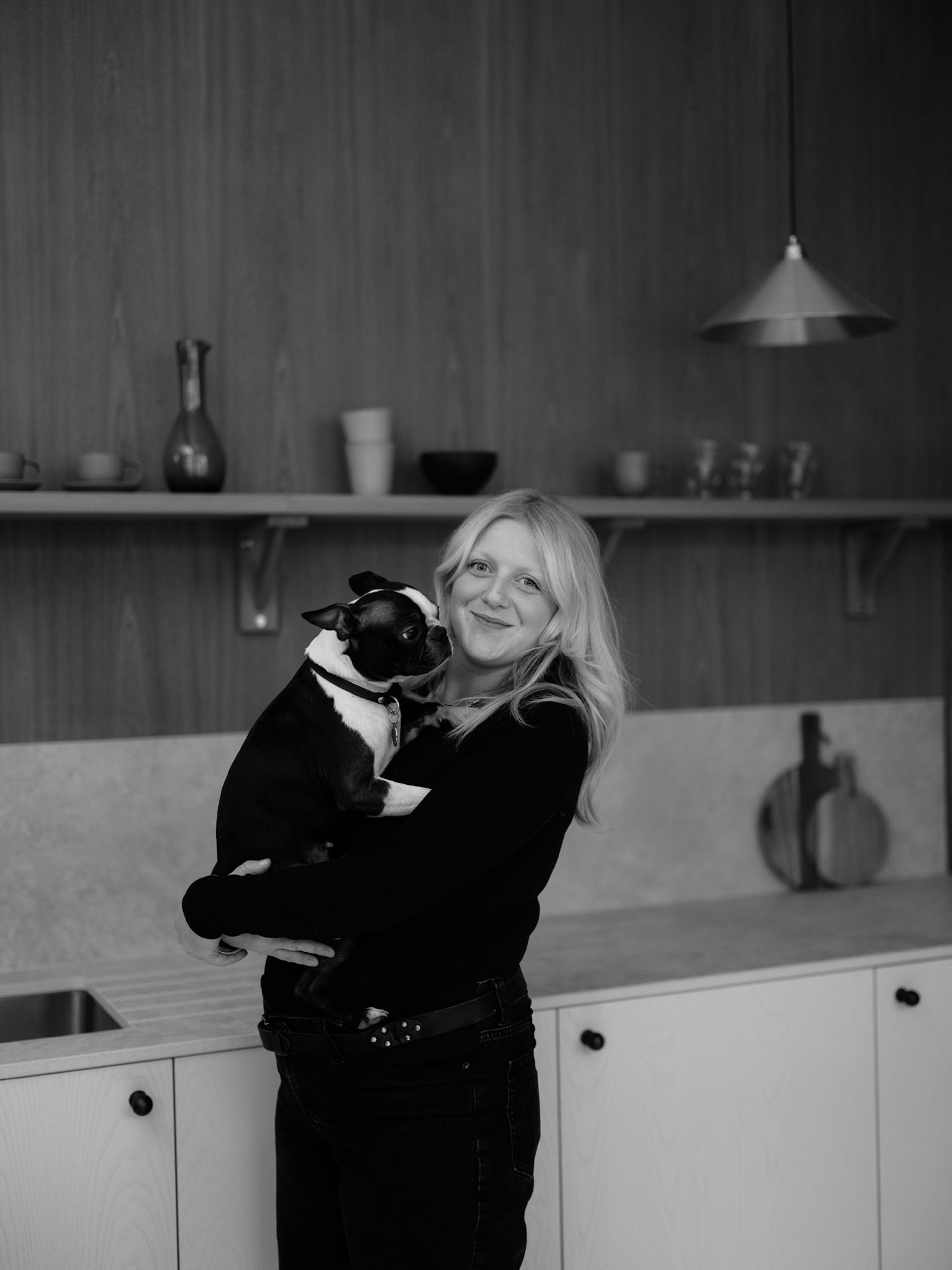 A black and white headshot of Freya Gibbons, a young lady with blonde hair standing in a kitchen wearing black and holding a dog while smiling at the camera
