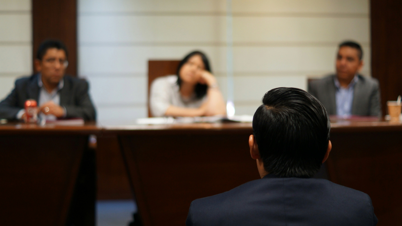 A man with a dark suit sits with his back to the camera in front of a table with three people sitting behind it