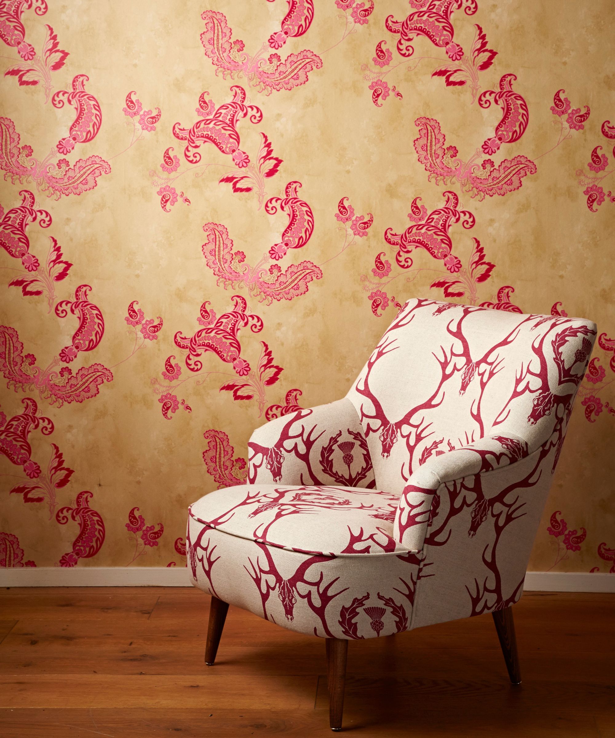 A patterned upholstered chair with a gold and pink patterned wallpapered wall behind. The floor is wooden.