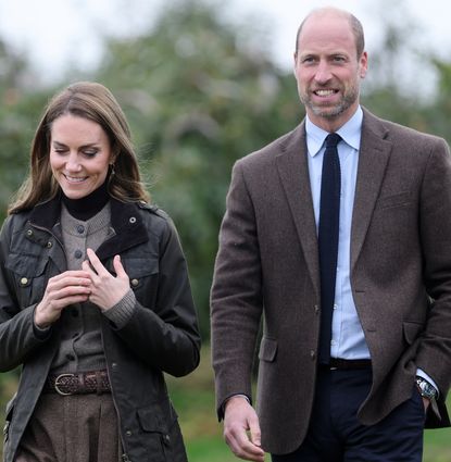 Prince William and Kate Middleton wearing brown and green jackets walking through a field