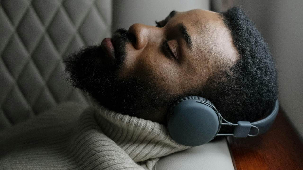 A man wearing headphones sleeping on the arm of a plane seat