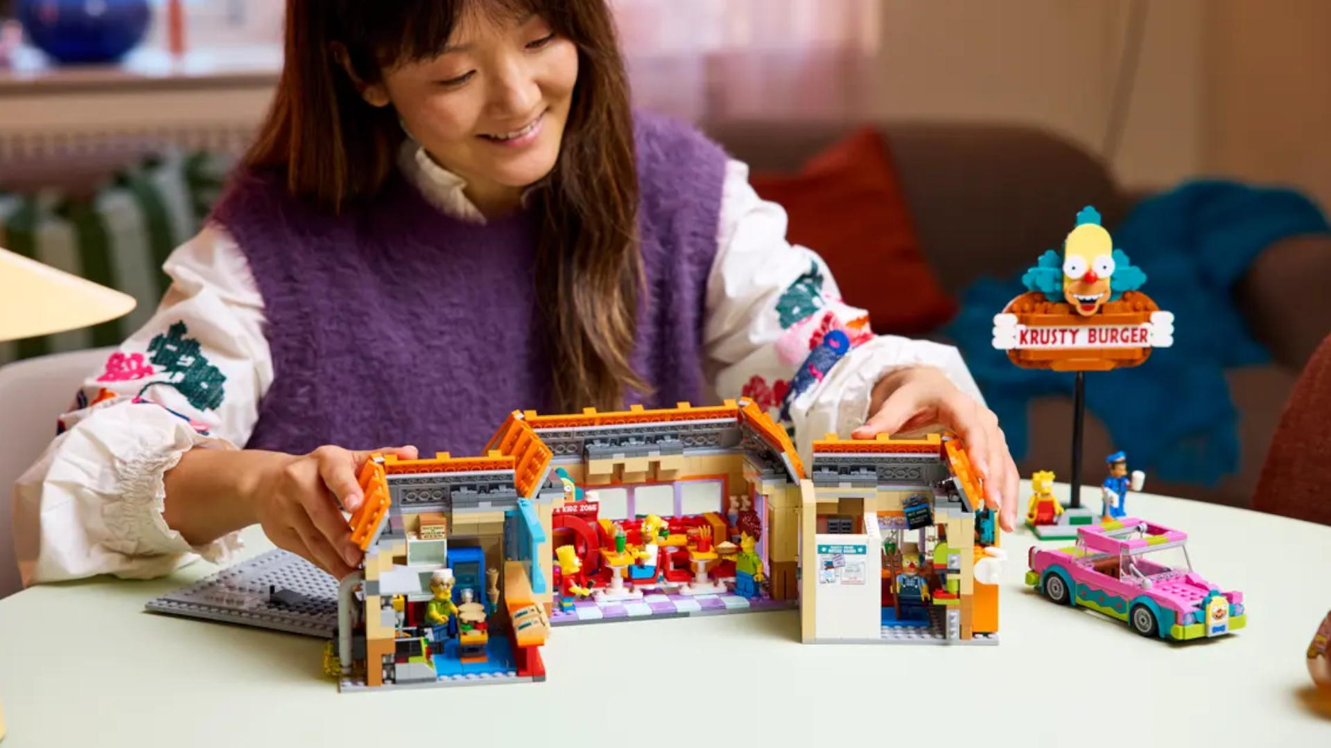 Lego Krusty Burger set on a plain surface, with a smiling woman opening the kit up